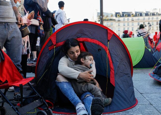 Mother cradling her child inside a small red tent at a crowded outdoor camp area