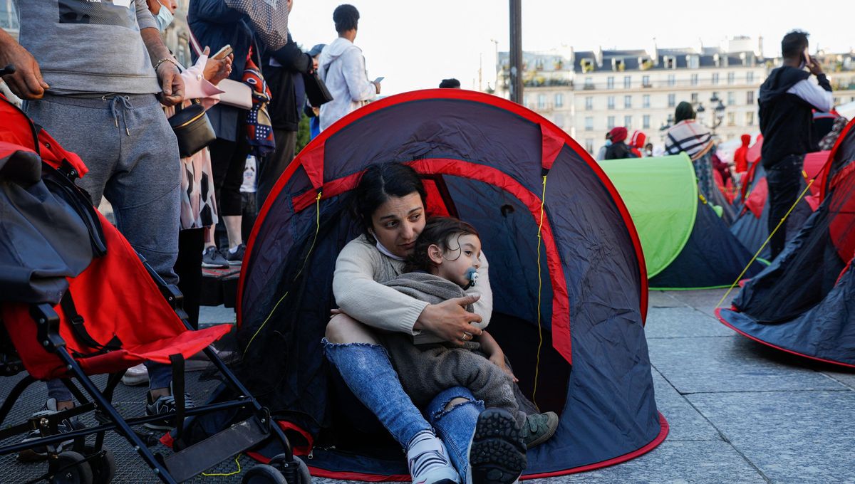 Mother cradling her child inside a small red tent at a crowded outdoor camp area