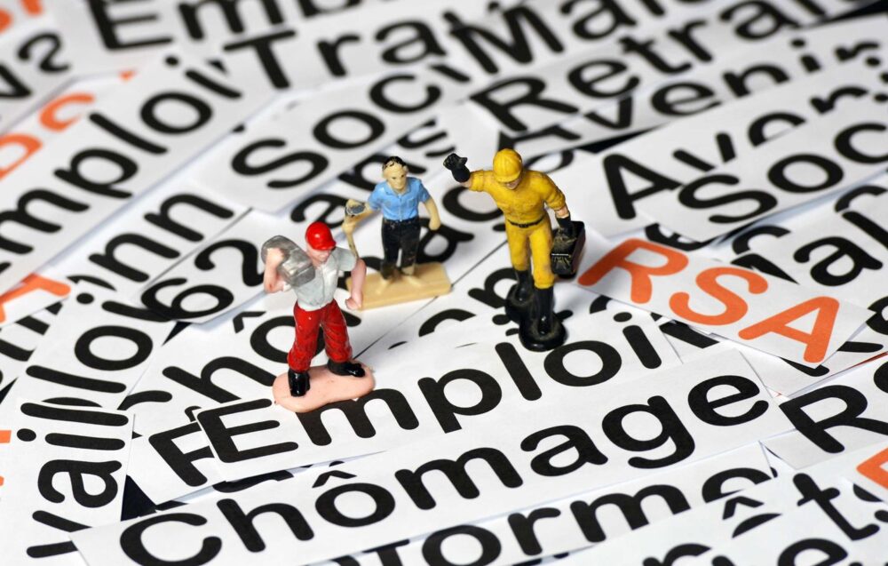 Miniature workers stand on scattered word cards about unemployment and employment (e.g., 'emploi', 'chômage'), illustrating labor market themes.