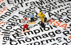 Miniature workers stand on scattered word cards about unemployment and employment (e.g., 'emploi', 'chômage'), illustrating labor market themes.