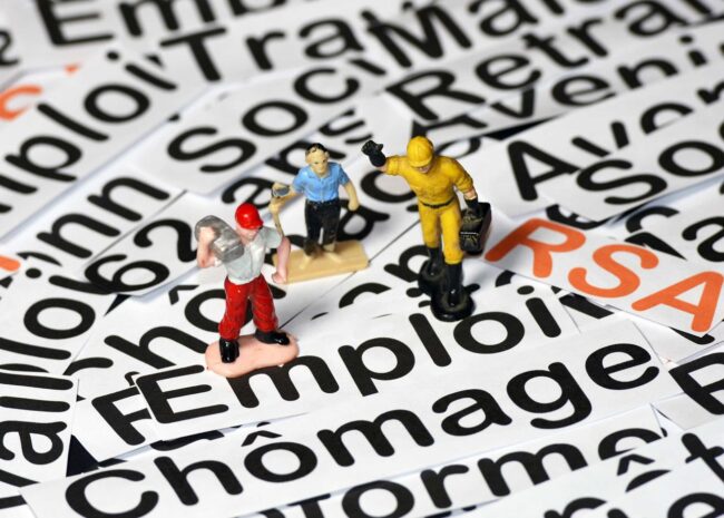 Miniature workers stand on scattered word cards about unemployment and employment (e.g., 'emploi', 'chômage'), illustrating labor market themes.