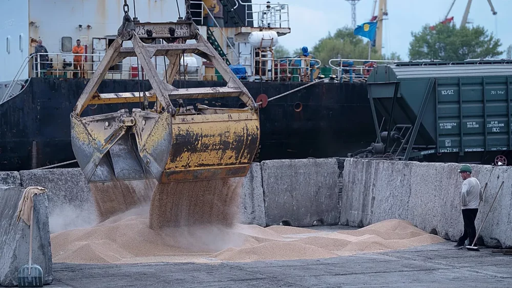 Industrial port scene with a large yellow excavator bucket dumping sand into a concrete pit as a worker watches from the right.