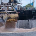 Industrial port scene with a large yellow excavator bucket dumping sand into a concrete pit as a worker watches from the right.