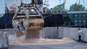Industrial port scene with a large yellow excavator bucket dumping sand into a concrete pit as a worker watches from the right.