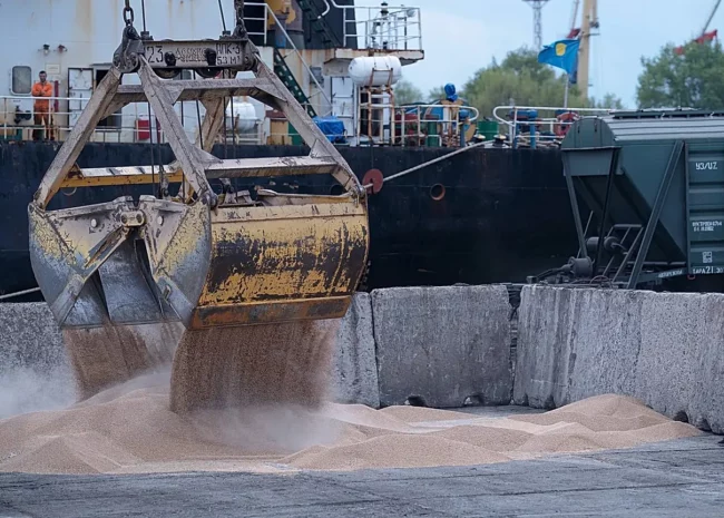Industrial port scene with a large yellow excavator bucket dumping sand into a concrete pit as a worker watches from the right.