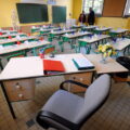 Teacher's desk in a bright yellow classroom, with a red binder, blue pencil cups, papers, and a vase of flowers; student desks arranged in rows behind.