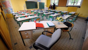 Teacher's desk in a bright yellow classroom, with a red binder, blue pencil cups, papers, and a vase of flowers; student desks arranged in rows behind.