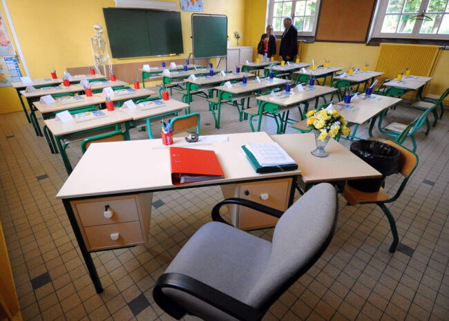 Teacher's desk in a bright yellow classroom, with a red binder, blue pencil cups, papers, and a vase of flowers; student desks arranged in rows behind.