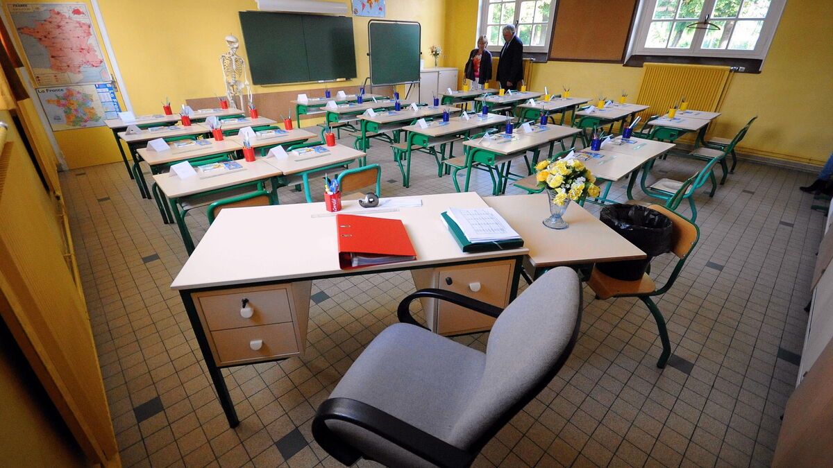 Teacher's desk in a bright yellow classroom, with a red binder, blue pencil cups, papers, and a vase of flowers; student desks arranged in rows behind.