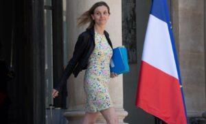 Woman in a floral dress with a black jacket walks past a stone column, carrying blue folders; a French flag is in the background.