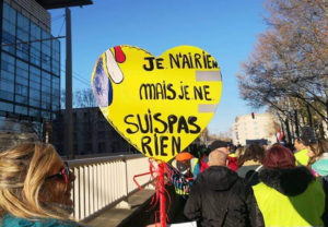 Protesters on a sunny street hold a large yellow heart-shaped sign with French words: 'Je n'ai rien mais je ne suis pas rien'.