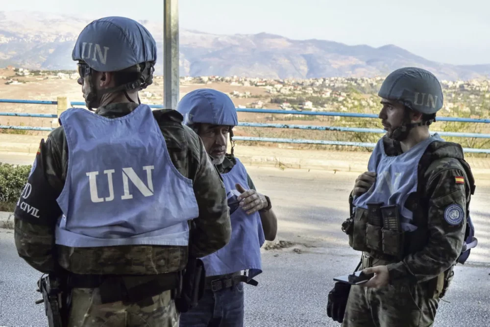 Three UN peacekeepers in blue vests and helmets standing and talking along a roadside, with a rural town and mountains in the background.