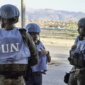 Three UN peacekeepers in blue vests and helmets standing and talking along a roadside, with a rural town and mountains in the background.