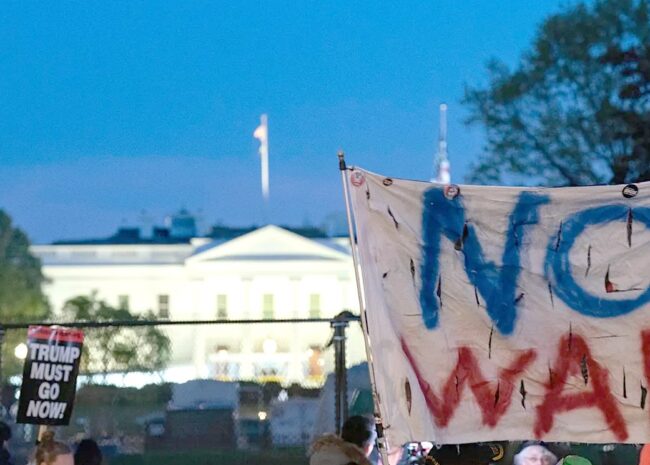 Protest at dusk with a torn banner reading NO WAR in blue and red; the White House lit in the background.