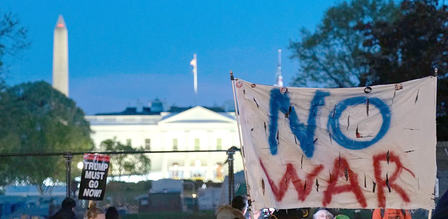 Protest at dusk with a torn banner reading NO WAR in blue and red; the White House lit in the background.