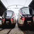 Two modern electric trains on parallel tracks at a station, facing the camera with overhead wires above them.