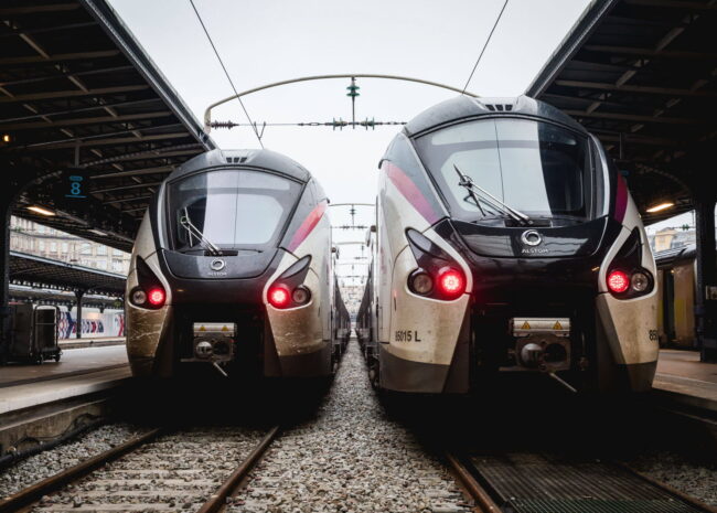 Two modern electric trains on parallel tracks at a station, facing the camera with overhead wires above them.