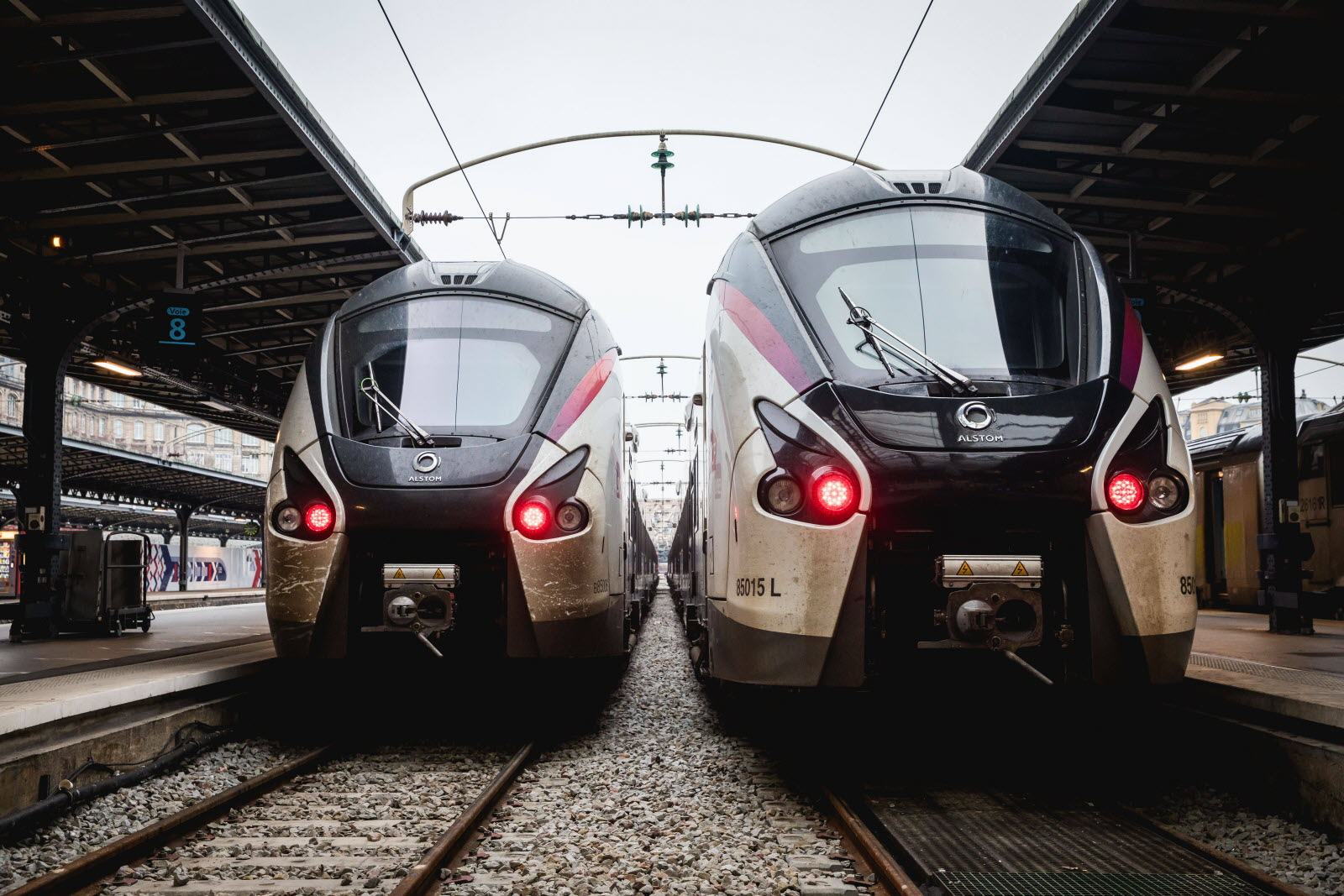 Two modern electric trains on parallel tracks at a station, facing the camera with overhead wires above them.