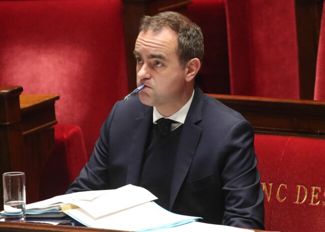 Man in a dark suit sits at a desk with papers, a pen in his mouth, in a red-velvet chamber.