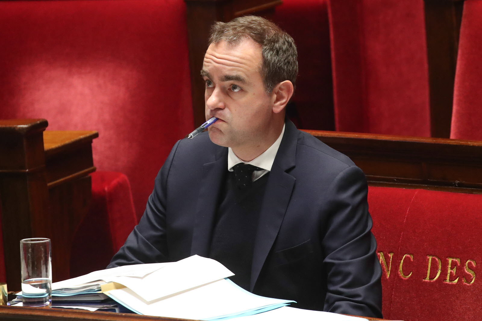 Man in a dark suit sits at a desk with papers, a pen in his mouth, in a red-velvet chamber.