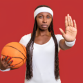 Female basketball player in a white jersey holds an orange basketball, raises her right hand toward the camera against a red background.