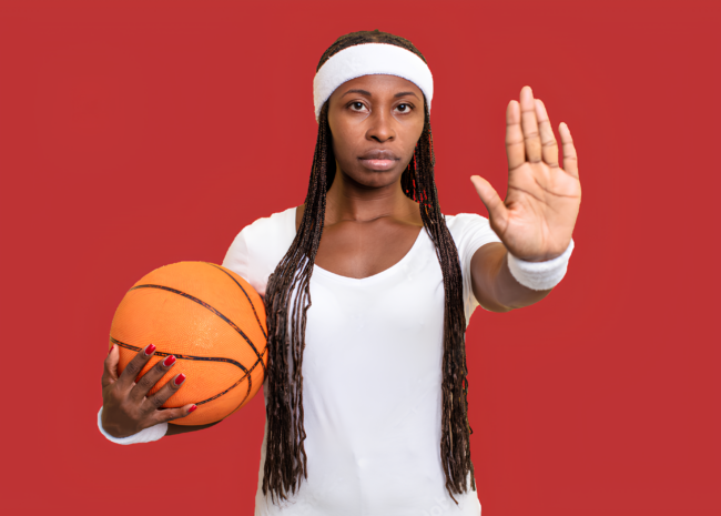 Female basketball player in a white jersey holds an orange basketball, raises her right hand toward the camera against a red background.