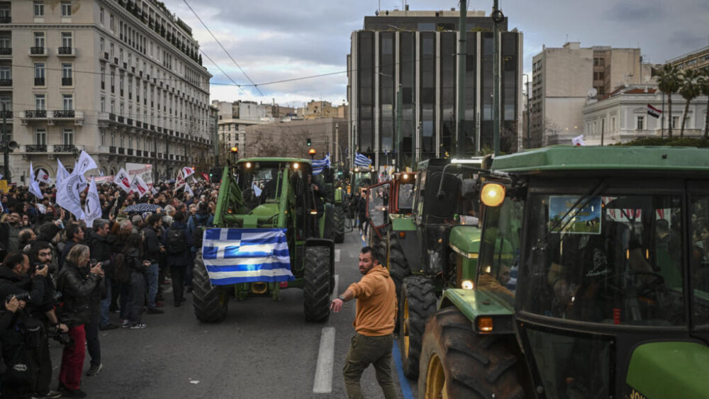 000-97F94ZQ - MultiPol 360 Tractor convoy protests in a city street with a Greek flag on the front tractor and a crowd of onlookers on the sidewalk.