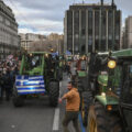 Tractor convoy protests in a city street with a Greek flag on the front tractor and a crowd of onlookers on the sidewalk.