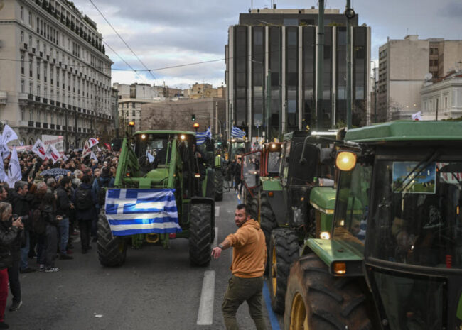 Tractor convoy protests in a city street with a Greek flag on the front tractor and a crowd of onlookers on the sidewalk.