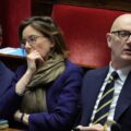 Three people in business attire sit in a parliamentary chamber with papers on the desk and a red upholstered bench behind them.