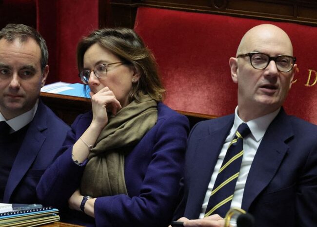Three people in business attire sit in a parliamentary chamber with papers on the desk and a red upholstered bench behind them.