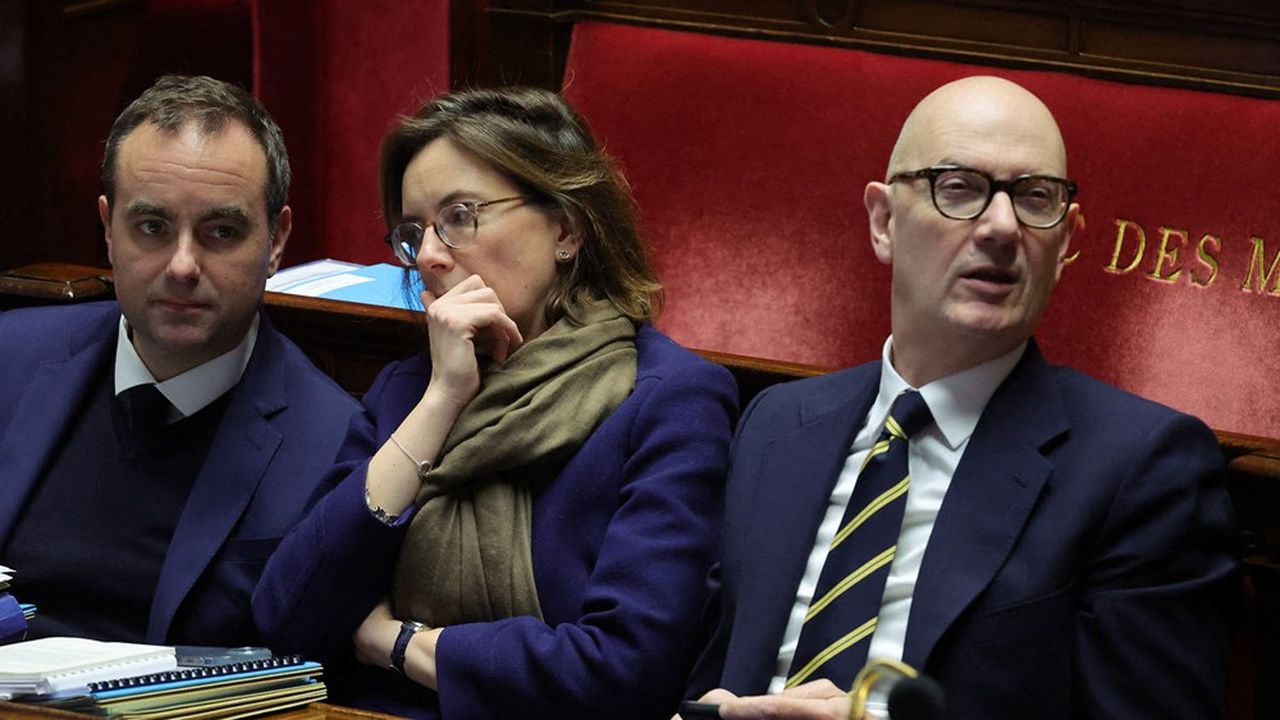 Three people in business attire sit in a parliamentary chamber with papers on the desk and a red upholstered bench behind them.