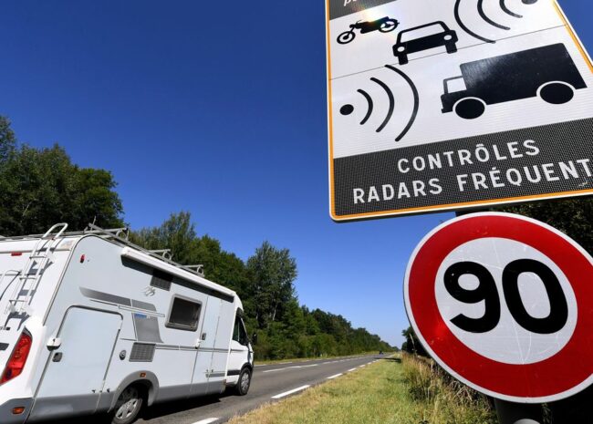 A white motorhome parked on the roadside with a large 90 km/h speed limit sign and a radar control sign in a rural area under a clear blue sky.