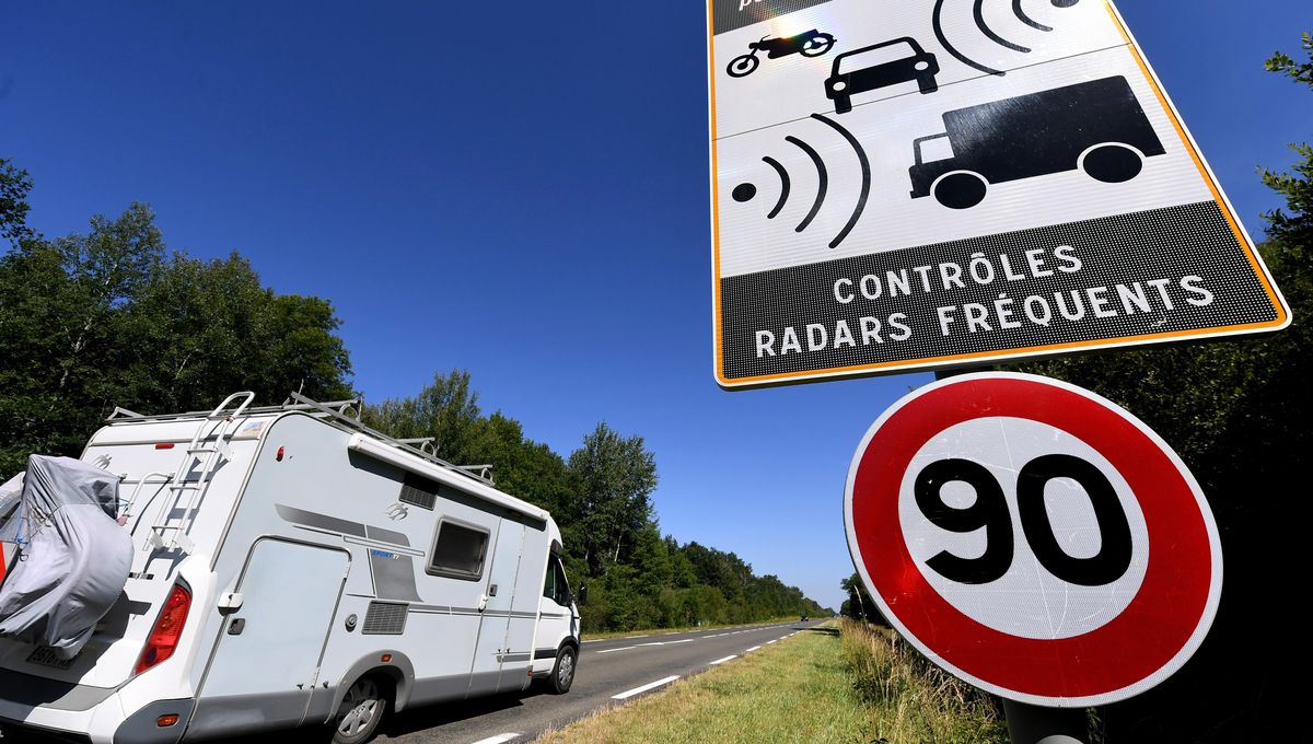 A white motorhome parked on the roadside with a large 90 km/h speed limit sign and a radar control sign in a rural area under a clear blue sky.