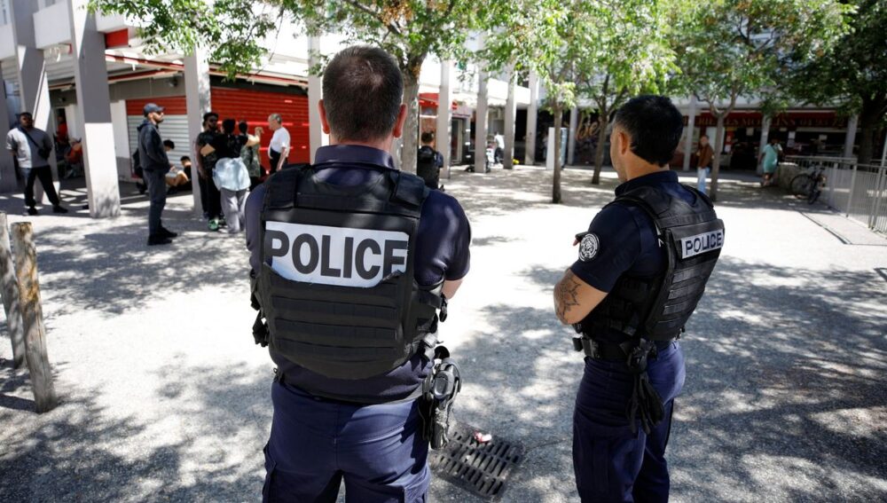 Two police officers in tactical vests stand with their backs to the camera in a sunny urban square, watching a group of people nearby.
