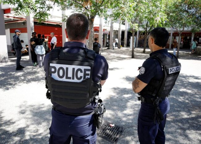 Two police officers in tactical vests stand with their backs to the camera in a sunny urban square, watching a group of people nearby.