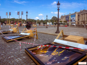 Outdoor plaza with large exhibition posters displayed on wooden frames, surrounded by red-and-white tape and pedestrians under a blue sky.