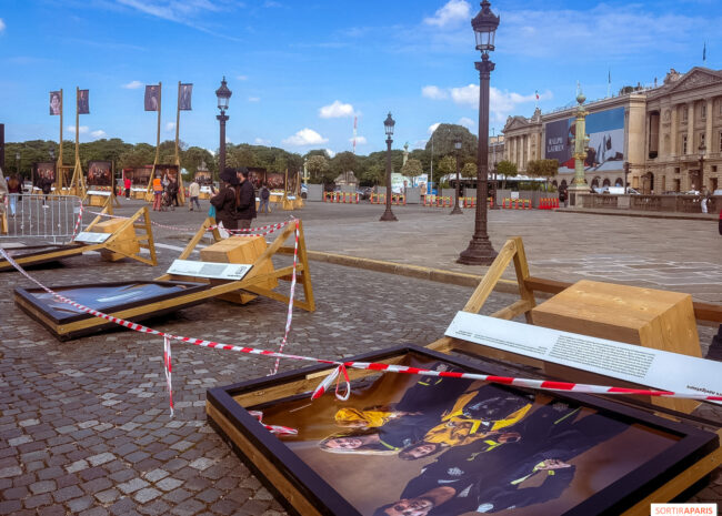 Outdoor plaza with large exhibition posters displayed on wooden frames, surrounded by red-and-white tape and pedestrians under a blue sky.