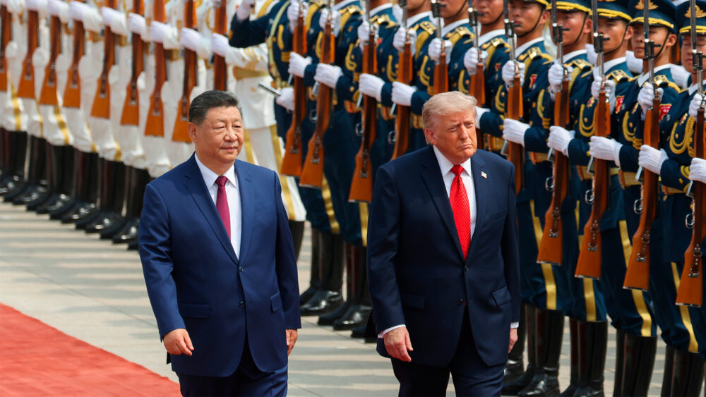 Two men in dark suits walk along a red carpet past a line of uniformed honor guards with rifles held upright.