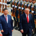 Two men in dark suits walk along a red carpet past a line of uniformed honor guards with rifles held upright.