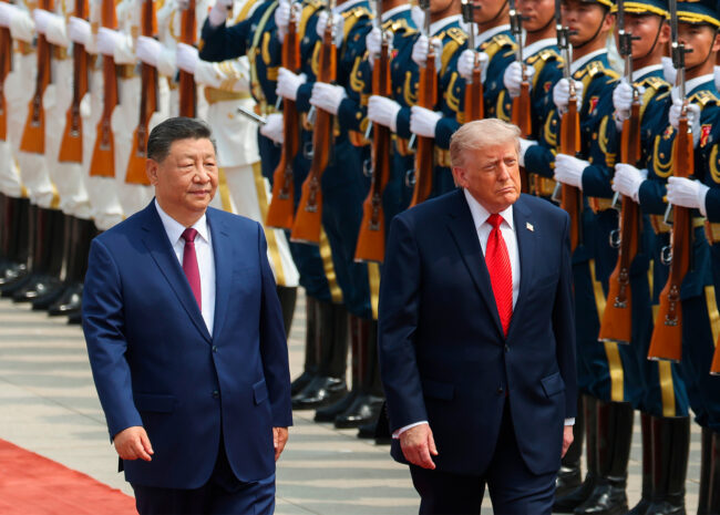 Two men in dark suits walk along a red carpet past a line of uniformed honor guards with rifles held upright.