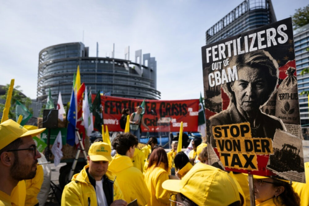 Protest scene with people in yellow jackets holding banners; a large poster reads 'FERTILIZERS OUT OF CBAM' and 'STOP VON DER TAX' in front of a modern urban building.