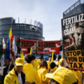 Protest scene with people in yellow jackets holding banners; a large poster reads 'FERTILIZERS OUT OF CBAM' and 'STOP VON DER TAX' in front of a modern urban building.