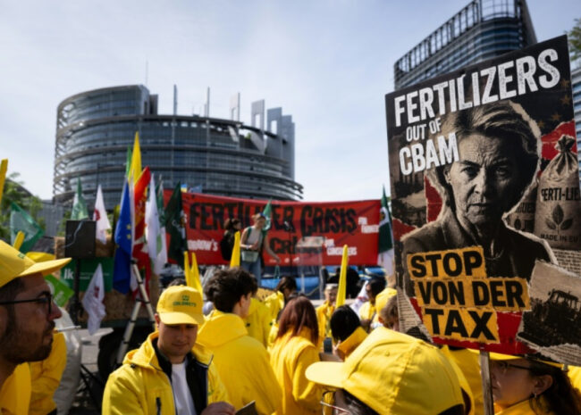 Protest scene with people in yellow jackets holding banners; a large poster reads 'FERTILIZERS OUT OF CBAM' and 'STOP VON DER TAX' in front of a modern urban building.