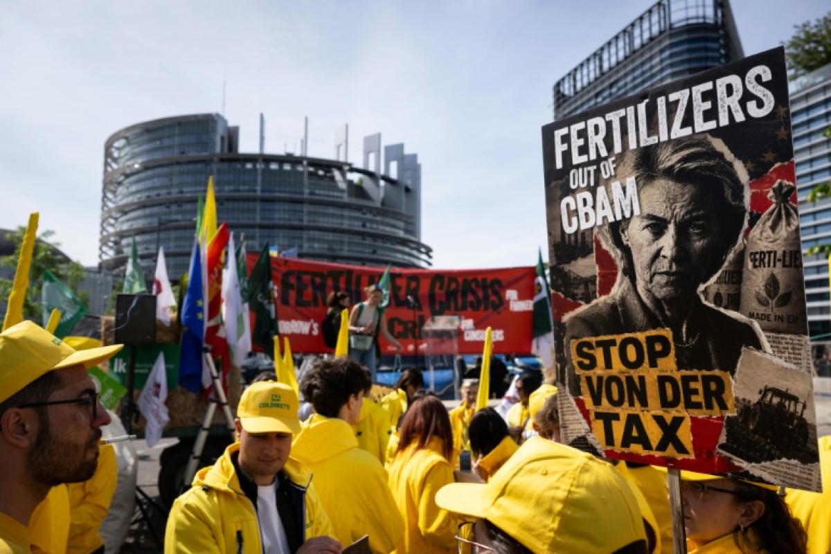 Protest scene with people in yellow jackets holding banners; a large poster reads 'FERTILIZERS OUT OF CBAM' and 'STOP VON DER TAX' in front of a modern urban building.