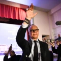 Man in a dark suit raises his right hand on stage during a press conference, with microphones and reporters visible nearby and a red curtain in the background.