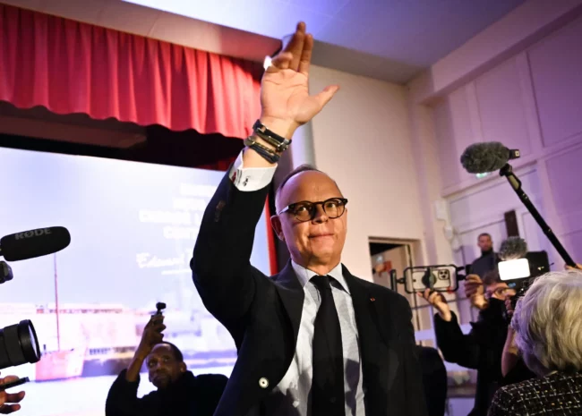 Man in a dark suit raises his right hand on stage during a press conference, with microphones and reporters visible nearby and a red curtain in the background.