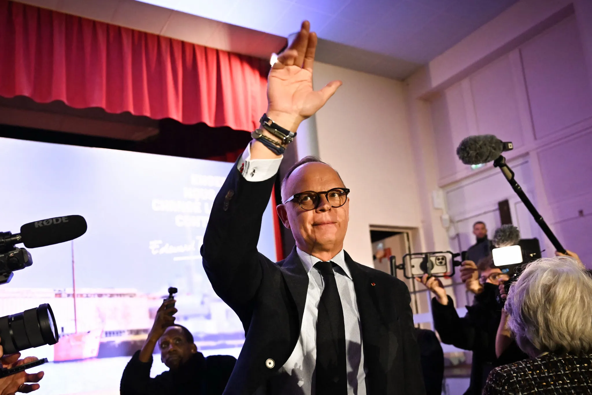 Man in a dark suit raises his right hand on stage during a press conference, with microphones and reporters visible nearby and a red curtain in the background.