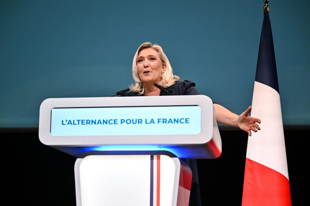 Female speaker at a podium delivering a speech; screen reads "L'Alternance Pour La France" with a French flag beside the stage.