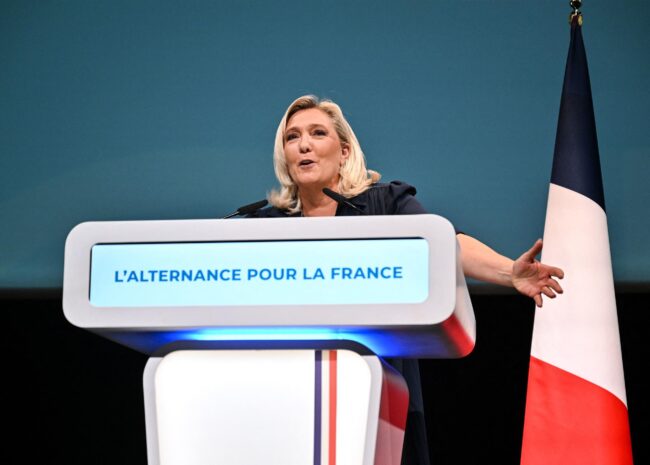 Female speaker at a podium delivering a speech; screen reads "L'Alternance Pour La France" with a French flag beside the stage.